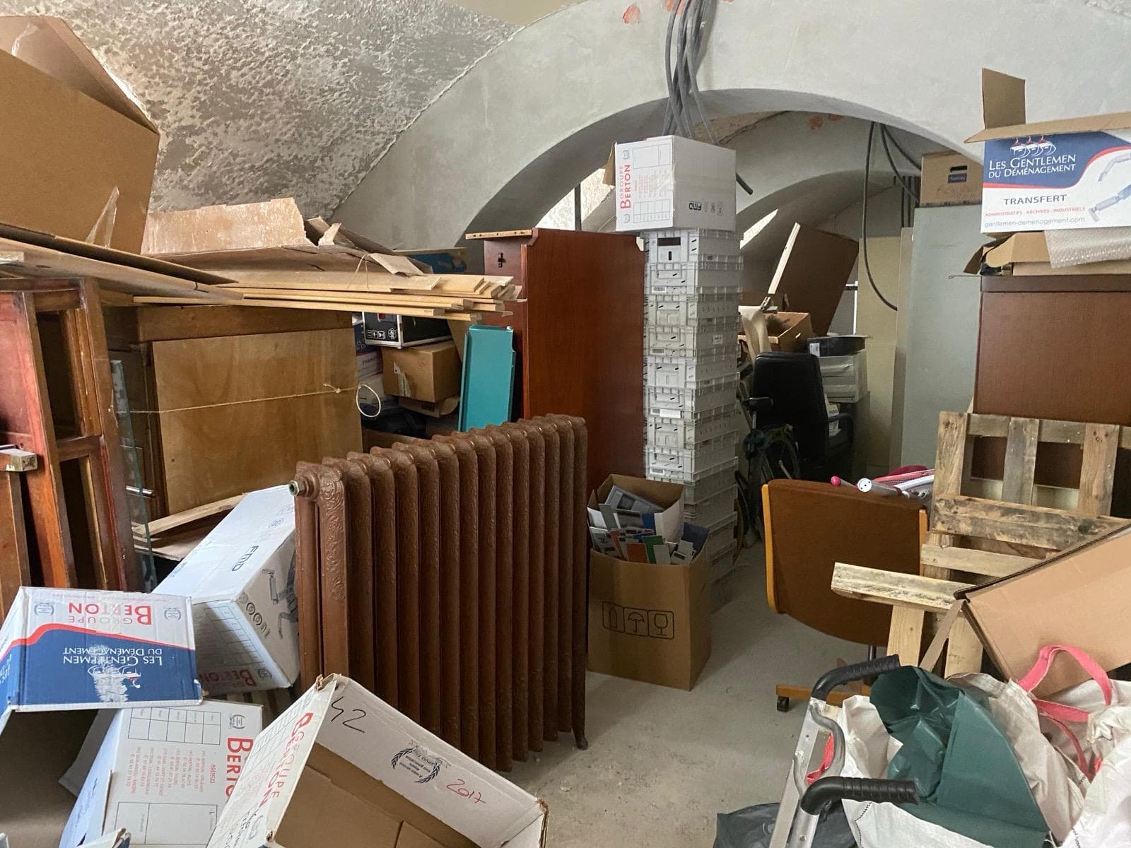 Cave voutée encombrée de radiateurs, cartons et meubles avant débarras